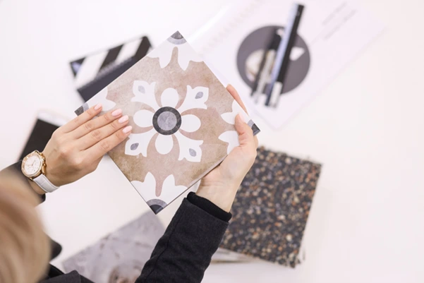 A woman holding a decorative ceramic tile with a floral pattern over a selection of other tile samples.